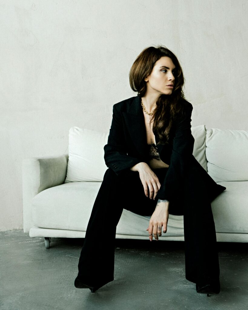 Stylish woman in a black suit sitting on a white sofa in a minimalistic interior with a neutral background.